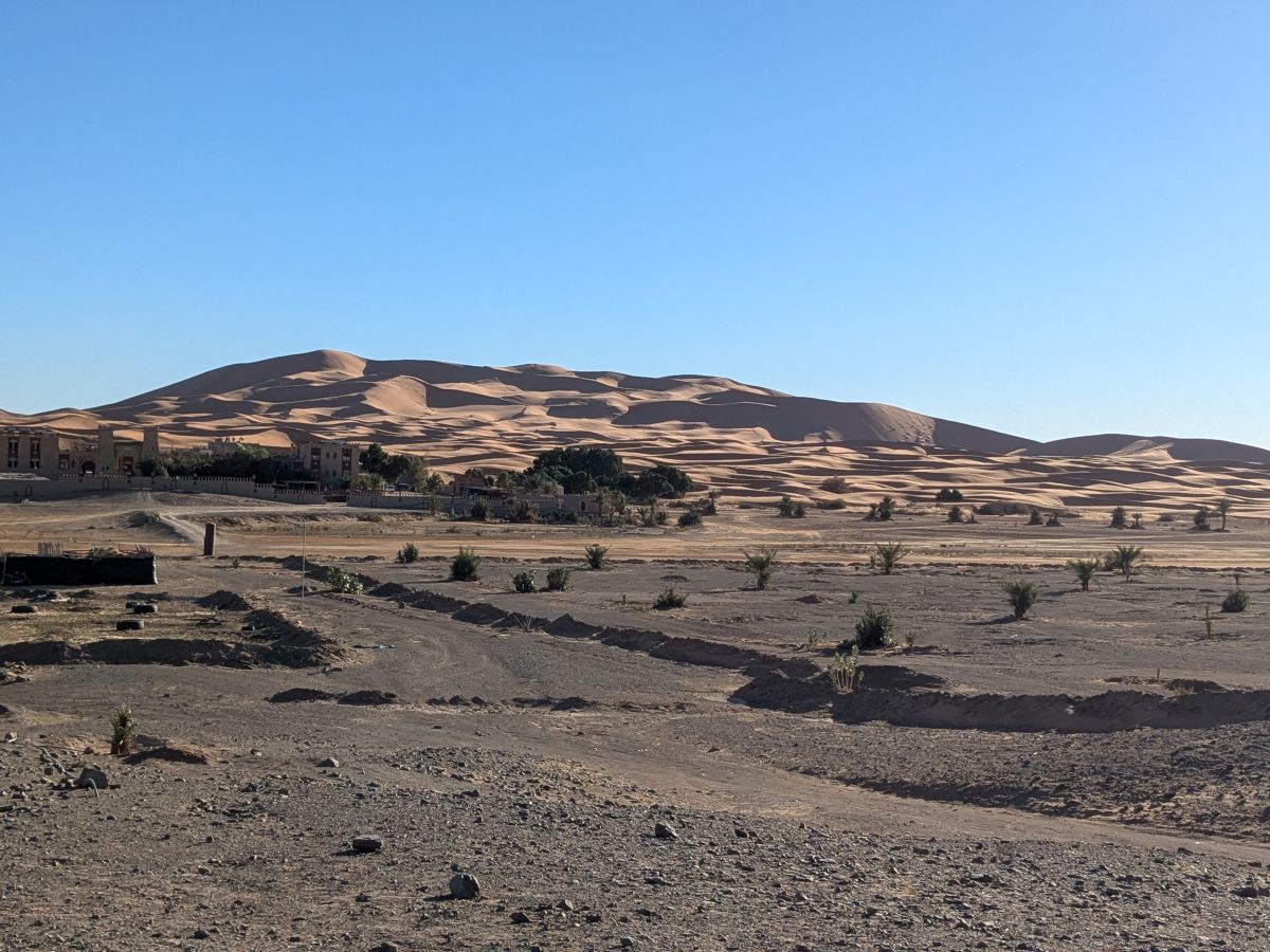 Looking out over a rocky desert landscape towards a walled resort complex at the foot of tall sand dunes.