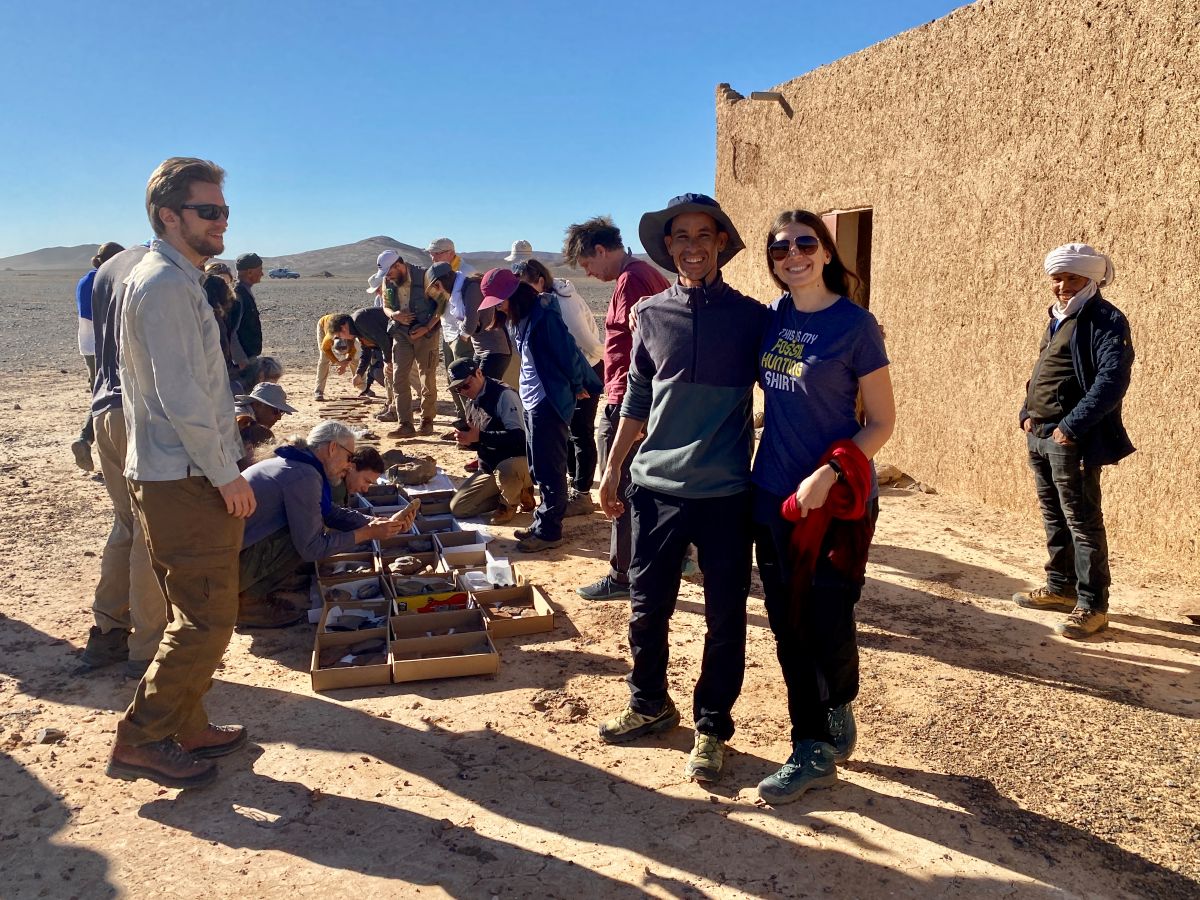 A group of conference attendees, two of whom are posing together for the photo, outside of a stone house. Many are looking at a wide selection of fossils displayed in boxes on the ground.