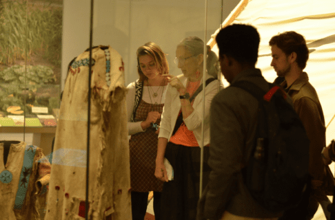 Three young people and an older adult wearing an orange shirt standing together looking into a case displaying a hide dress.