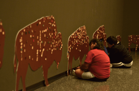 Two children sit on the floor in front of bison cut-outs of varying sizes displayed on the wall in front of them. The bison are each decorated with small visitor-placed tiles.