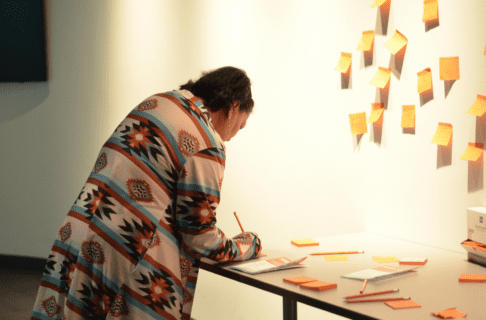An individual stands over a table, writing something on an orange sticky note to put up with others on the wall in front of them.