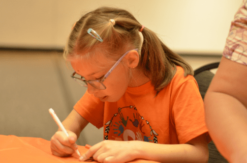 A child wearing an orange shirt sits at a table with an orange table cloth and writes something on a small wooden tile with a marker.