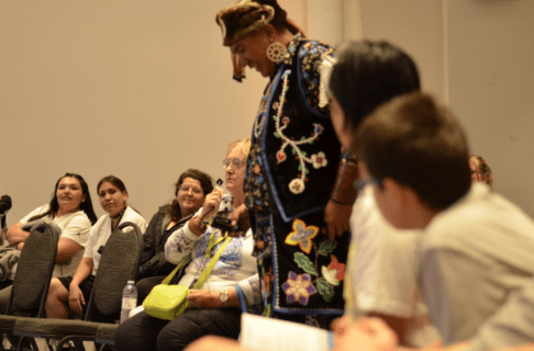 An individual in Indigenous regalia walks through a seated crowd.