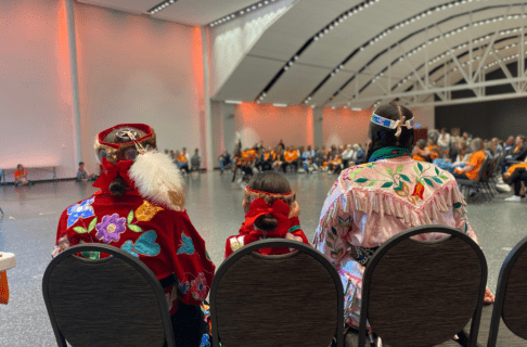 From behind, seated, two adults and a child wearing traditional regalia facing an open area where a dancer performs in front of a seated crowd, many of whom wear orange shirts.