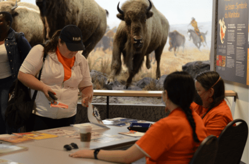 Two individuals wearing orange t-shirts seated at a table as they engage with a visitor in an orange shirt and white sweater.