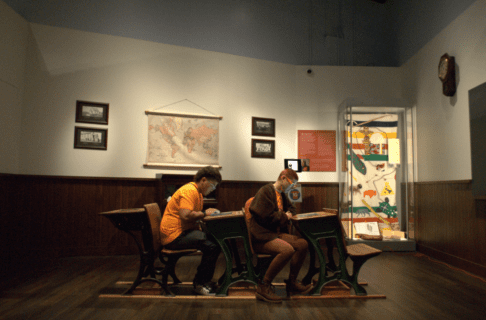 Two young adults wearing orange t-shirts sit at desks in the Schoolhouse exhibit, engaging with digital material embedded in the desk tops.