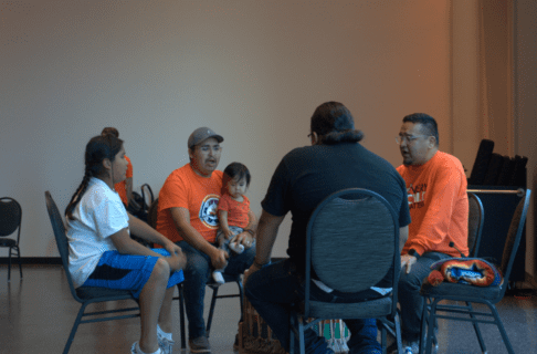 Four individuals seated in a circle, one of whom has a toddler on their knee, with a drum between them as they sing together.