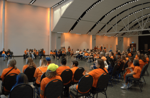 A crowd seated in a semi-circle, most of whom are wearing orange shirts.