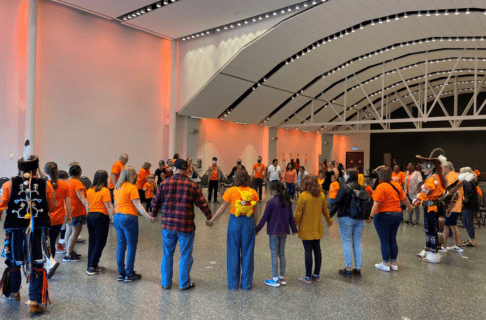A group of people, most of whom are wearing orange shirts, standing and holding hands in a large circle.