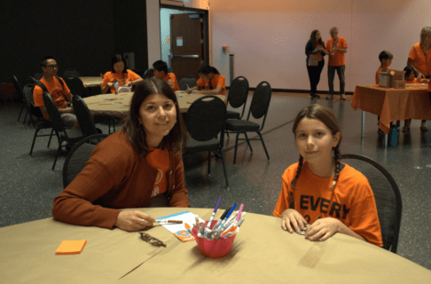 An adult and a child seated at a round table, both wearing orange shirts, smiling for a photo as they decorate small wooden tiles.