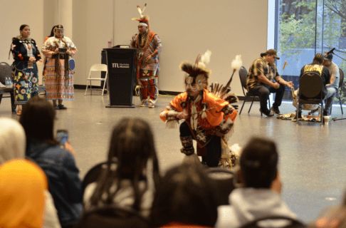 Over the heads of a crowd looking towards a dancer in Indigenous regalia with other dancers watching from the side.