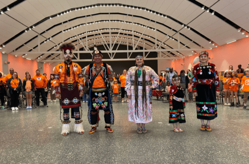 Five dancers in regalia, four adults and one child, stand in a row smiling for a photo with a crowd of orange shirt-wearing individuals behind them.