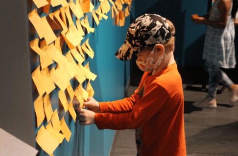 A child wearing an orange shirt and a baseball cap places an orange sticky note onto a wall next to many more sticky notes.