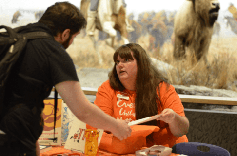 A woman wearing an orange t-shirt seated at a table with event materials assists a visitor.