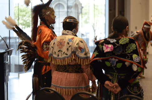From behind, three Indigenous dancers in full regalia standing together to the side as another person dances in the background.