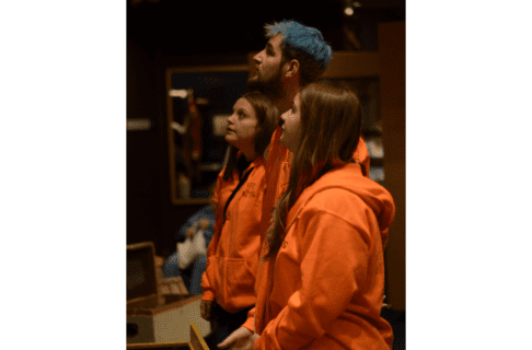 Three individuals from the side, standing side by side and looking up at something out of frame. All three wear orange shirts of sweaters.