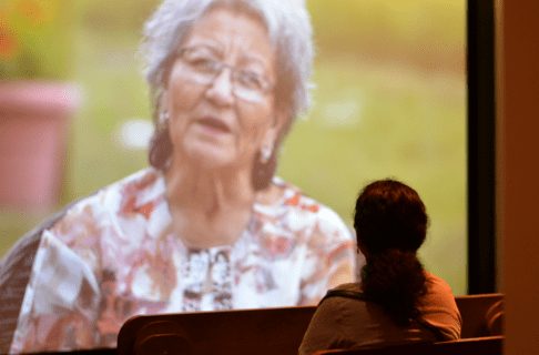 An individual from behind as they sit and watch a video projected on a large screen, showing an older woman outdoors speaking to camera.