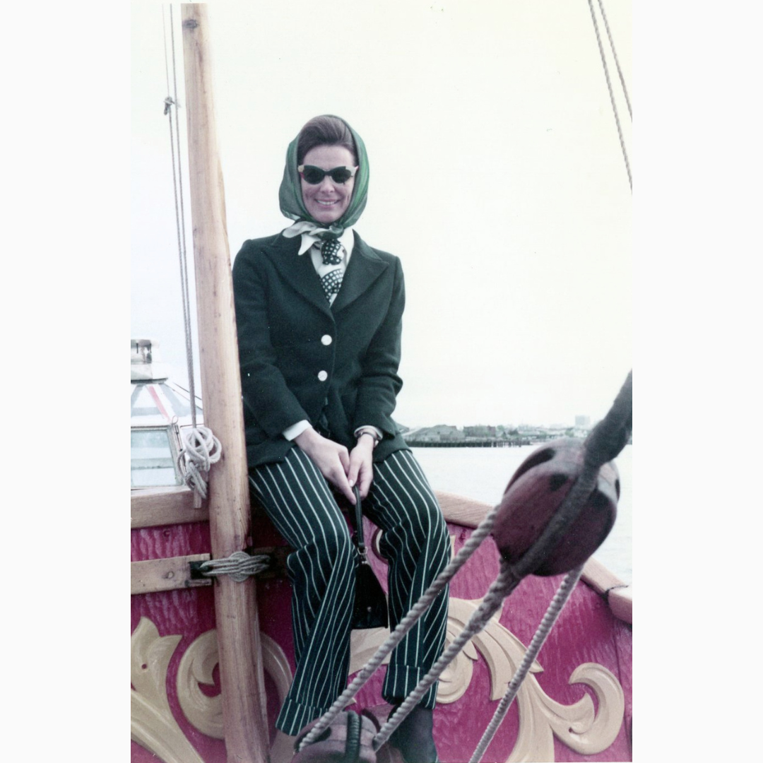 Young Tannis Richardson sits on the deck of the Nonsuch replica off the coast of British Columbia (Victoria, BC 1972), wearing a dark coat, headscarf, sunglasses, and striped trousers, with rigging and water visible behind.