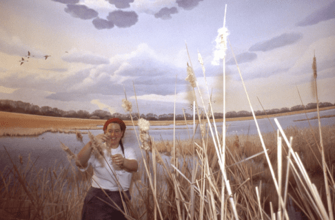 A smiling woman standing in a partially constructed marsh diorama placing bulrushes.