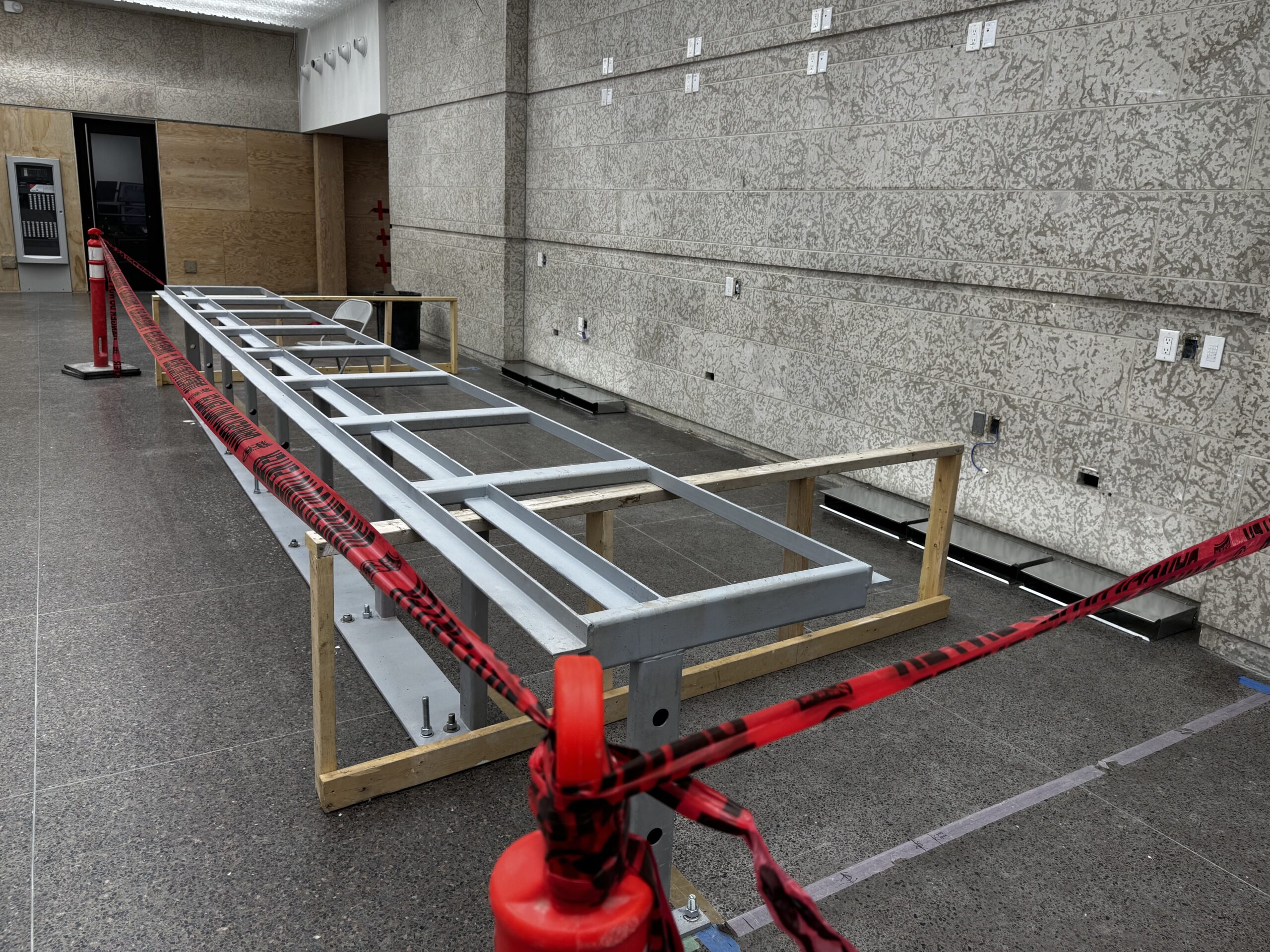Metal framework of a long low desk in front of a stone wall in the Museum Foyer. Red caution tape creates a perimeter.