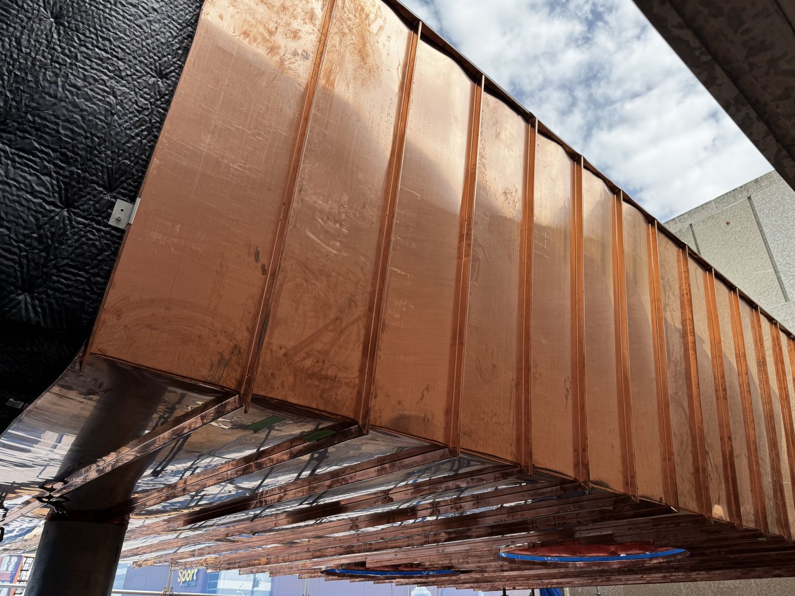 Shining copper cladding lining the side of an awning under a lightly-clouded blue sky.