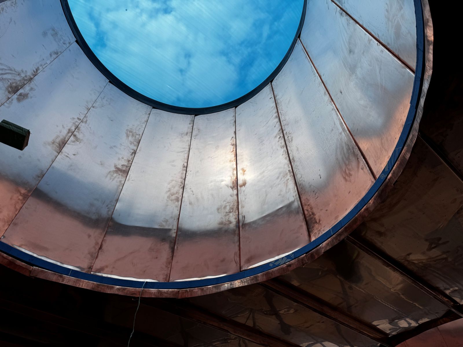 Looking up through a circular framed skylight to a lightly clouded sky through a transparent blue material. The skylight is lined with copper cladding around the interior.