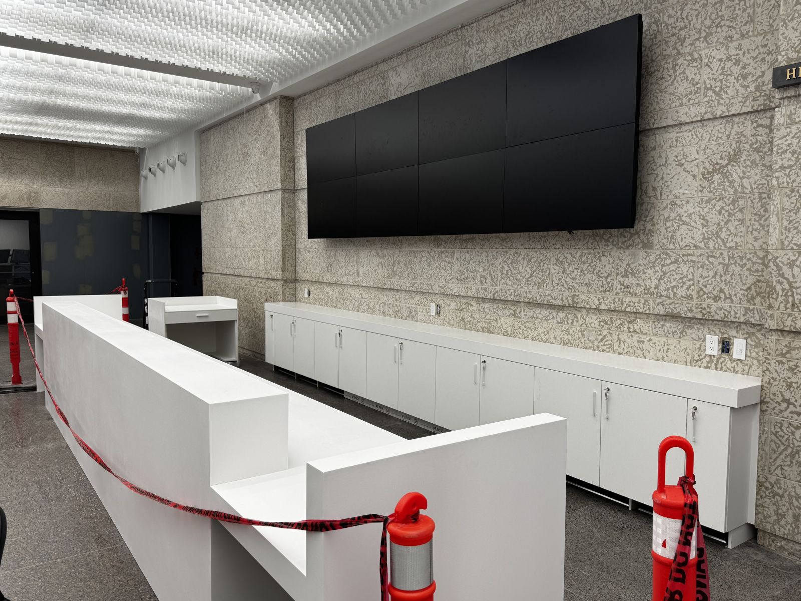 A white-clad long, low desk in front of a stone wall with white shelving units at the base in the Museum Foyer. Above the shelving units is a panel of eight digital screens.