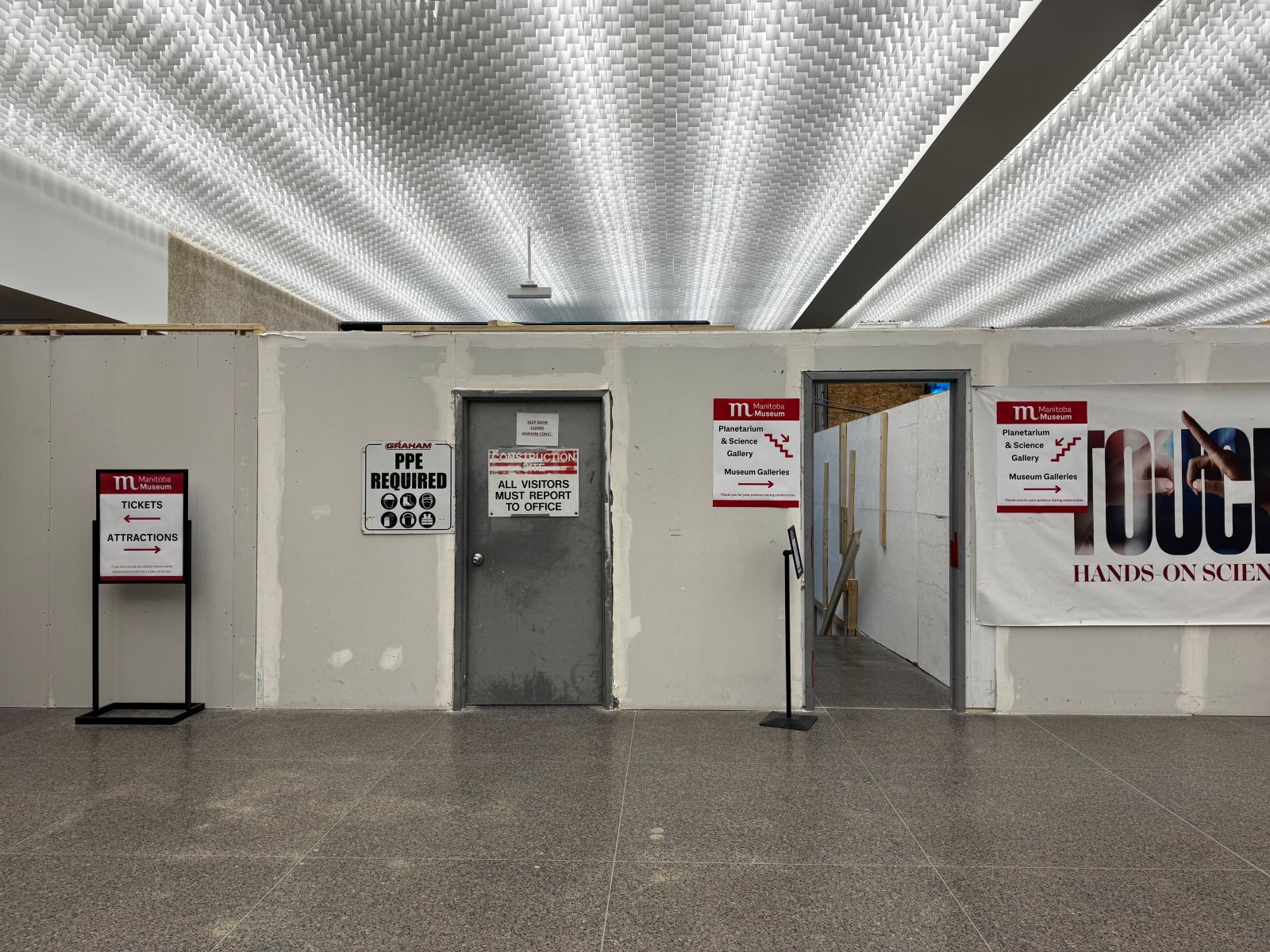 Construction hoarding wall with two steel framed fire doors. One door, to the active construction zone is closed, while the other goes down to the open stairwell to reach the lower level of the Museum. Signage on the hoarding directs visitors in the direct of the stairwell or the Box Office.