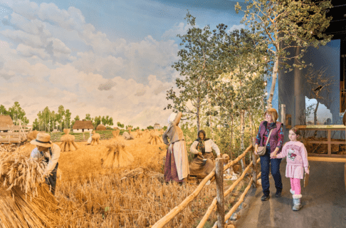An adult and child walk hand in hand down a ramped walkway next to a life size museum diorama depicting a farming family during a rye harvest.