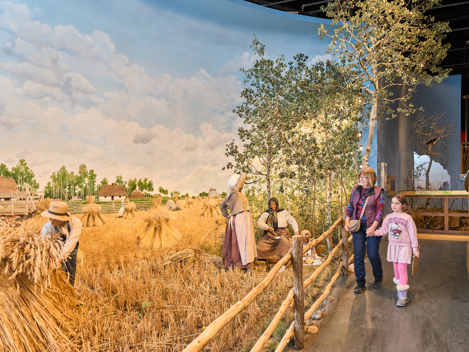 An adult and child walk hand in hand down a ramped walkway next to a life size museum diorama depicting a farming family during a rye harvest.