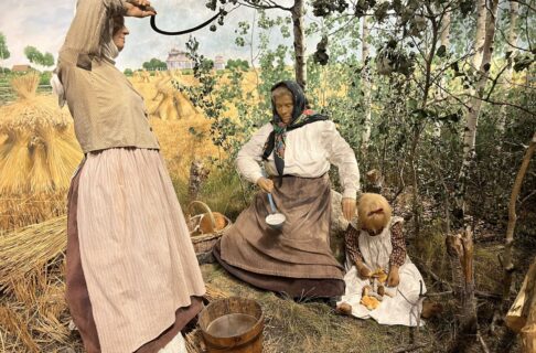 Rye farm diorama. Three mannequins depicting a standing adult woman wiping her brow while holding a sickle, standing next to a seated older woman lifting a ladle from a pail of water and a child seated on the group with a selection of mushrooms in her lap.