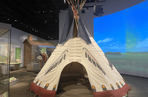A full-sized tipi set up in the centre of a Museum Gallery with a digital mural of a open grassy plain behind it.