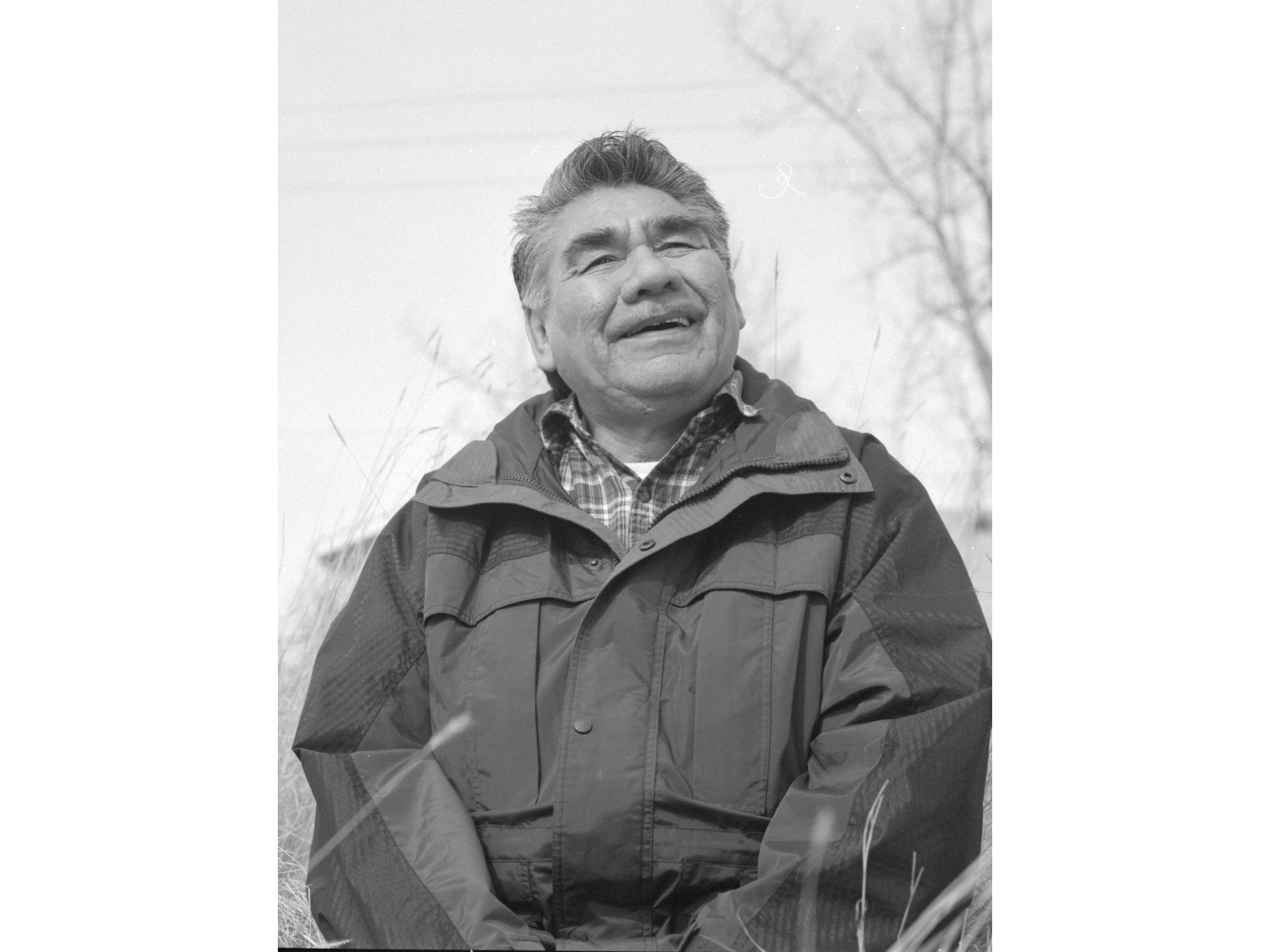 A black and white photo of a man from the waist up, wearing a windbreaker jacket and sitting outside, smiling over and past the camera.
