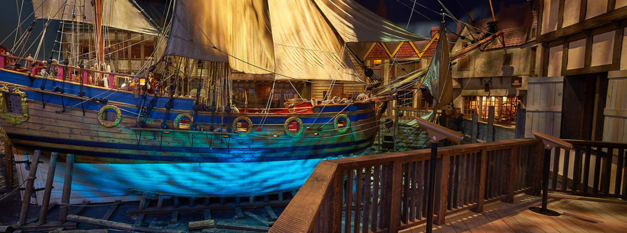 A view of the Nonsuch ketch, a full-sized replica, from a pier-like exhibit space. The ship is dry-docked and you can see the white paint of the hull, as well as the blue stripes and carvings around the portholes along the side. There are two masts with the sails hanging down.