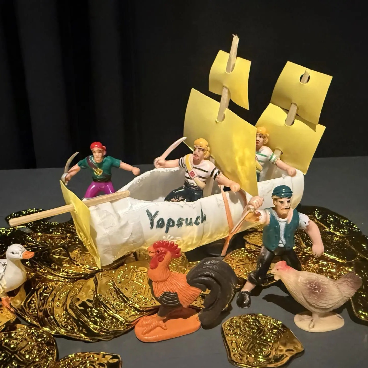 A two-mast sailing ship labelled as the Yopsuch made out of a plastic yogurt bottle with skewer and paper sails. Plastic pirate figures surround the vessel on a pile of plastic coins.
