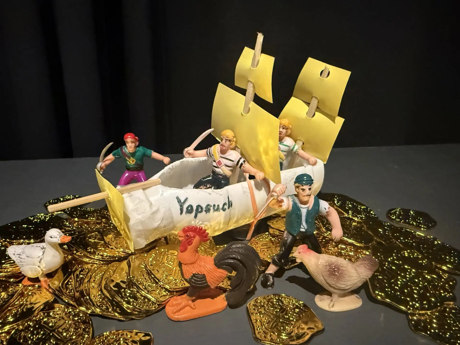 A two-mast sailing ship labelled as the Yopsuch made out of a plastic yogurt bottle with skewer and paper sails. Plastic pirate figures surround the vessel on a pile of plastic coins.