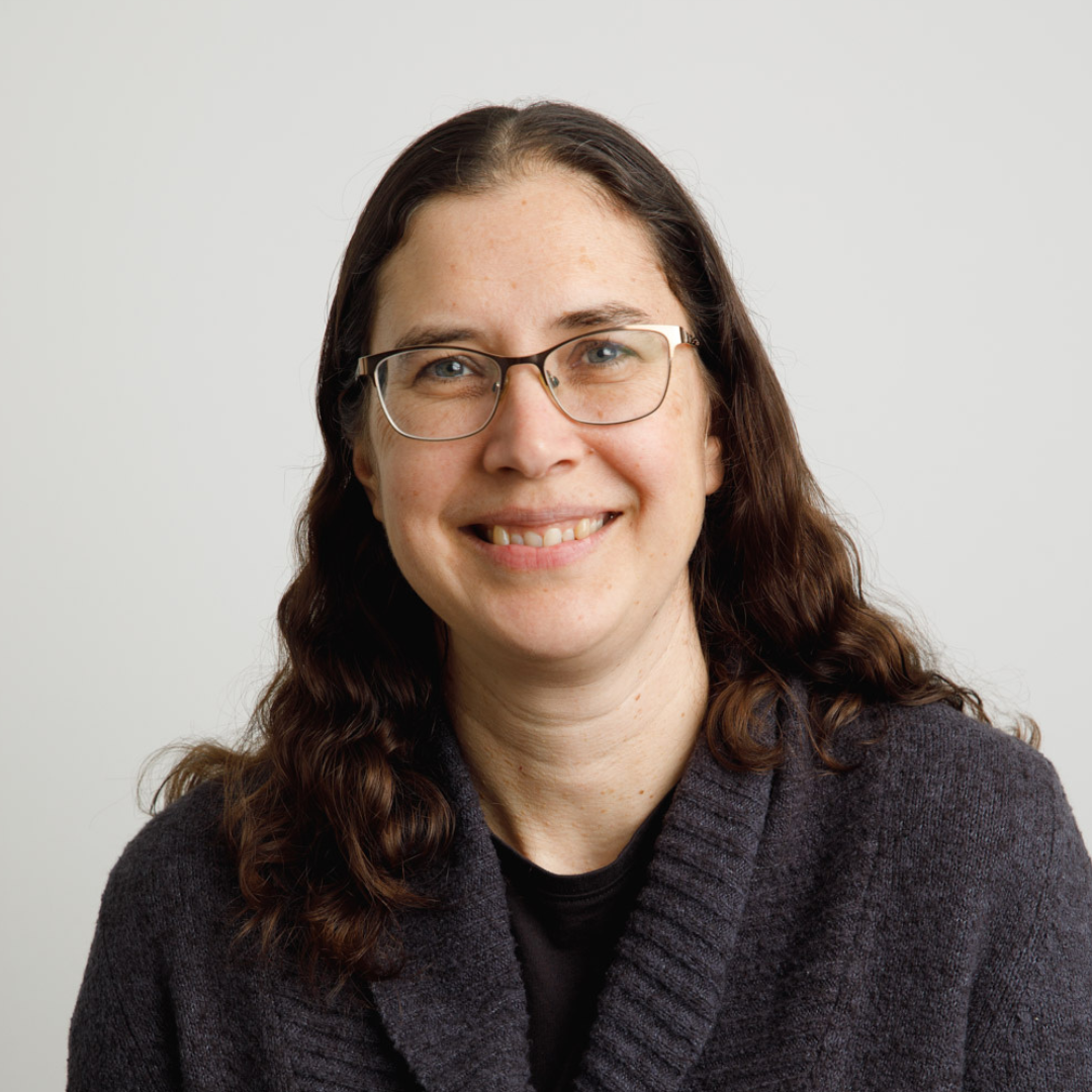 Formal headshot of Sonya Hauri-Thiessen, a smiling woman wearing a dark-coloured knit sweater, with wavy brunette hair going past her shoulders, and wire-rimmed glasses.