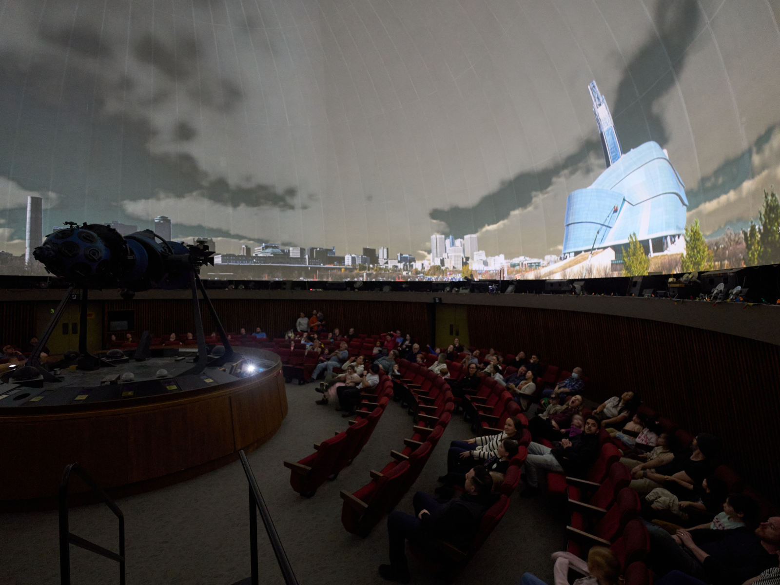 Photo of the interior of the Planetarium dome with audience members seated encircling the Zeiss star projector and a nighttime Winnipeg skyline projected on the ceiling dome.