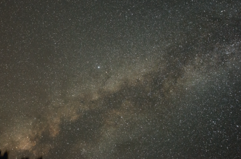 Photo taken looking up into the Milky Way from the ground below. A hazy band of light and stars stretching across the night sky.